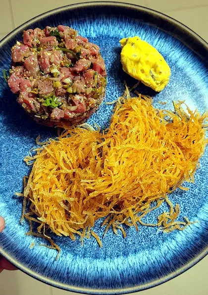 Beef tartare on a blue plate: hand-chopped raw beef with herbs, capers and mustard, a quenelle of bone-marrow sabayon, and a tangle of thin straw fries.