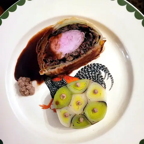 A slice of beef Wellington on a white plate — pink beef inside a dark mushroom layer and golden pastry — with a dark sauce and a disc of sliced leek.