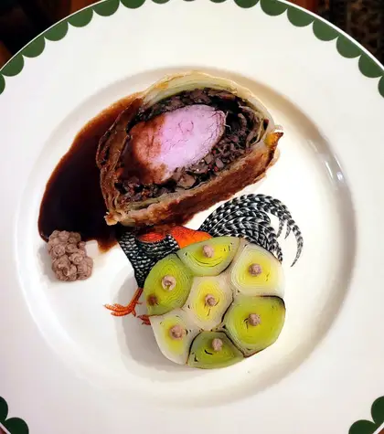 A slice of beef Wellington on a white plate — pink beef inside a dark mushroom layer and golden pastry — with a dark sauce and a disc of sliced leek.
