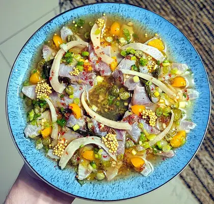 A blue-rimmed bowl of ceviche held in one hand: strips of white fish in a citrus marinade with red onion, spring onion, chilli and pieces of mango.