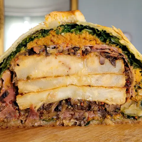 A vegetarian pithivier cut open on a wooden board, showing its layers of cabbage, butternut, mushroom duxelles and sliced celeriac inside the puff pastry.
