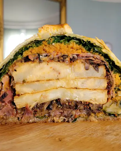 A vegetarian pithivier cut open on a wooden board, showing its layers of cabbage, butternut, mushroom duxelles and sliced celeriac inside the puff pastry.