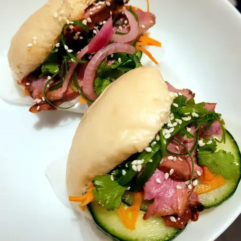 Two folded bao buns on a white plate, filled with sliced duck, pickled red onion, cucumber and coriander, sprinkled with sesame seeds.