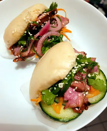 Two folded bao buns on a white plate, filled with sliced duck, pickled red onion, cucumber and coriander, sprinkled with sesame seeds.
