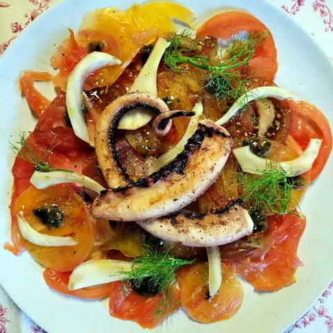 Tomato carpaccio on a pale plate, topped with charred octopus tentacles, shaved fennel, capers and fennel fronds.
