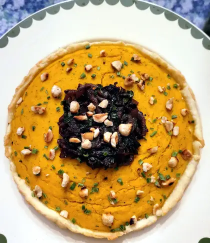 A pumpkin pie in a pale pastry case, its orange filling topped with dark caramelised onions, chopped hazelnuts and chives.
