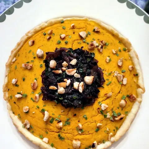 A pumpkin pie in a pale pastry case, its orange filling topped with dark caramelised onions, chopped hazelnuts and chives.