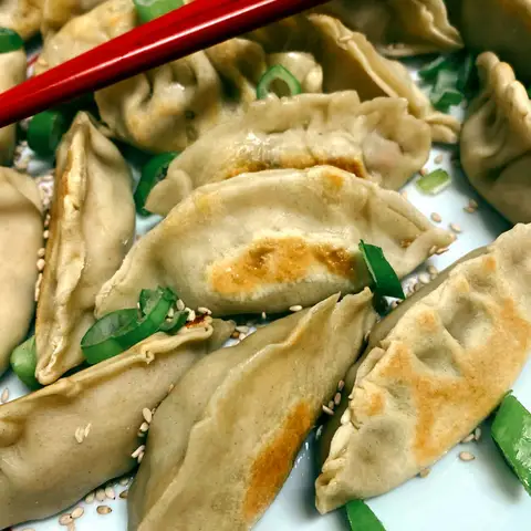 Pan-fried gyozas with golden-brown bases, scattered with spring onion and sesame seeds, a pair of red chopsticks laid across the plate.