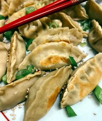 Pan-fried gyozas with golden-brown bases, scattered with spring onion and sesame seeds, a pair of red chopsticks laid across the plate.
