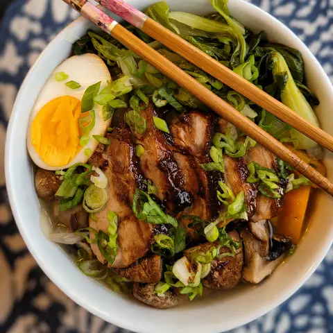 A bowl of ramen seen from above: slices of glazed pork belly, half a soft-boiled egg, spring onion, mushrooms and pak choi in broth, with chopsticks resting across the rim.