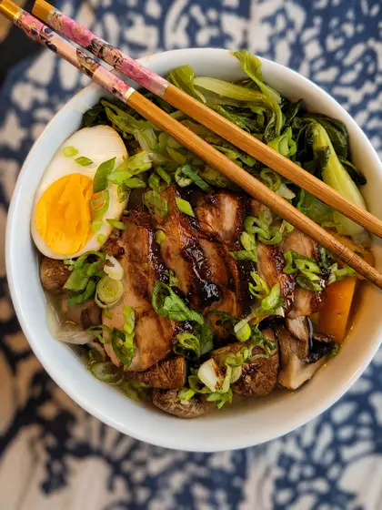 A bowl of ramen seen from above: slices of glazed pork belly, half a soft-boiled egg, spring onion, mushrooms and pak choi in broth, with chopsticks resting across the rim.