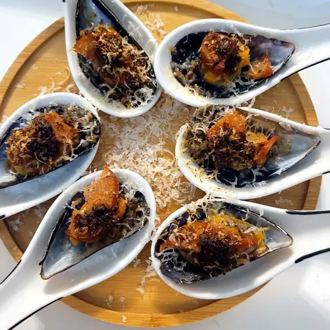 Six mussels on the half shell, each filled with chorizo and grated cheese and browned, arranged in a circle of white porcelain spoons on a wooden board.