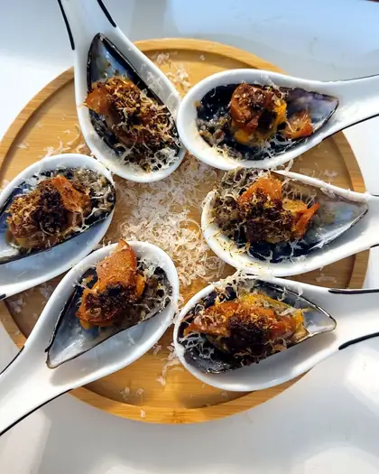 Six mussels on the half shell, each filled with chorizo and grated cheese and browned, arranged in a circle of white porcelain spoons on a wooden board.