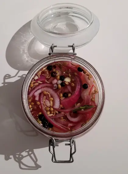 Looking straight down into an open glass jar of quick pickled red onion, sliced onion curled in a pink brine with coriander seeds, black peppercorns and a bay leaf.