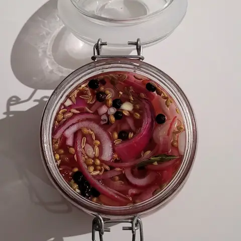 Looking straight down into an open glass jar of quick pickled red onion, sliced onion curled in a pink brine with coriander seeds, black peppercorns and a bay leaf.
