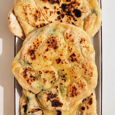 A stack of soft, golden naan flatbreads with lightly charred spots, served on a plate.