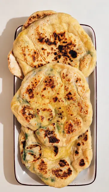 A stack of soft, golden naan flatbreads with lightly charred spots, served on a plate.
